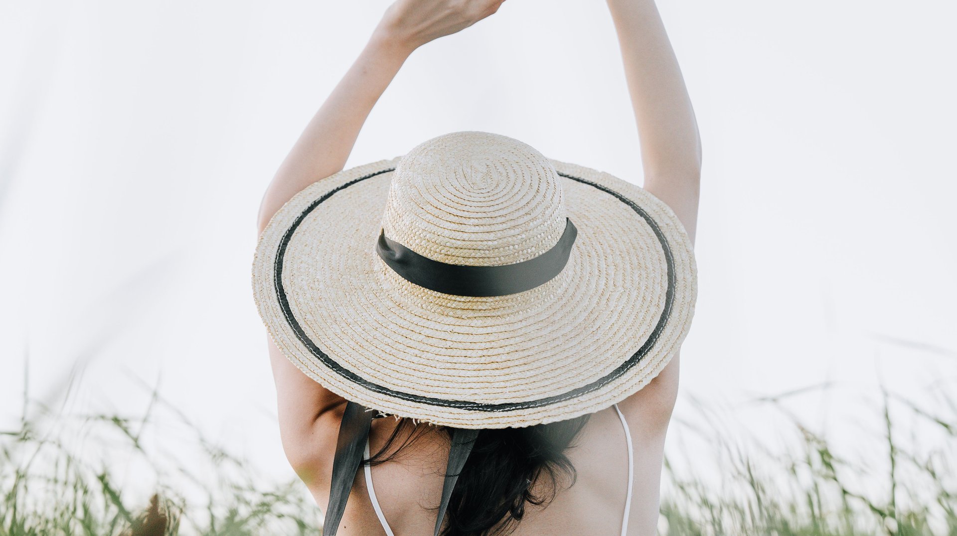 Back View of a Woman with Arms Raised in a Grassy Summer Field, Wearing a Sun Hat.
