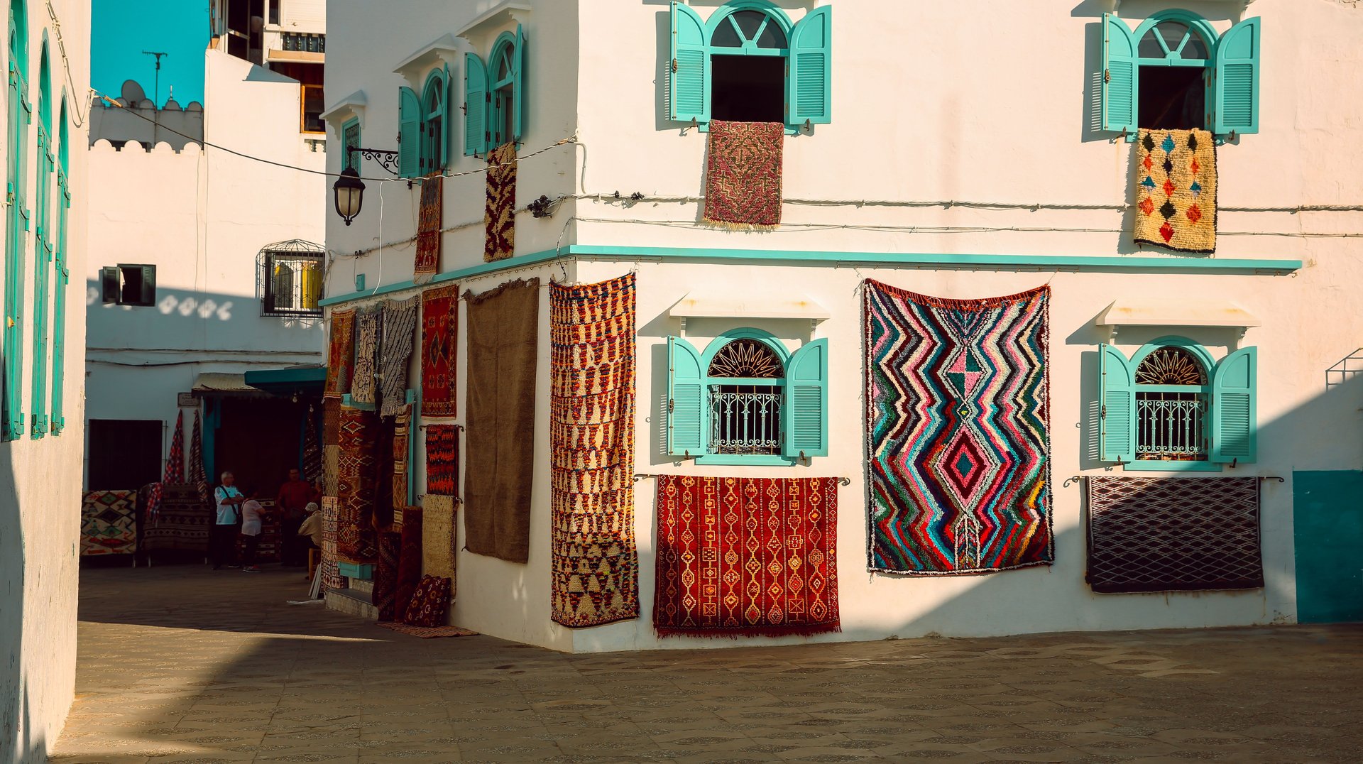 Vibrant Street in Asilah, Morocco Featuring Traditional Woven Rugs Hung on White Buildings with Turquoise Shutters.