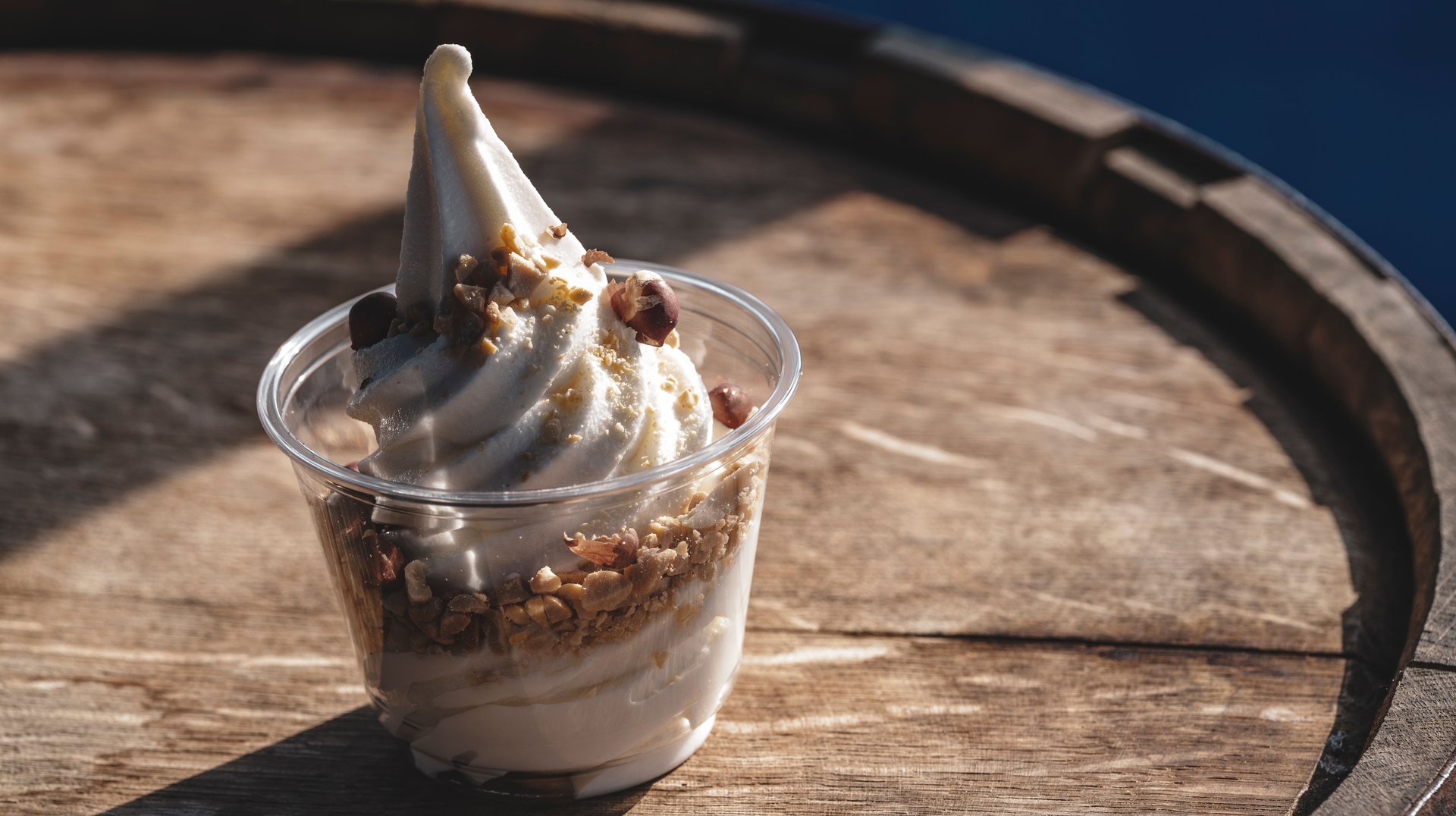 a Cup of Soft Serve Ice Cream with Toppings on a Wooden Barrel in Sunlight.