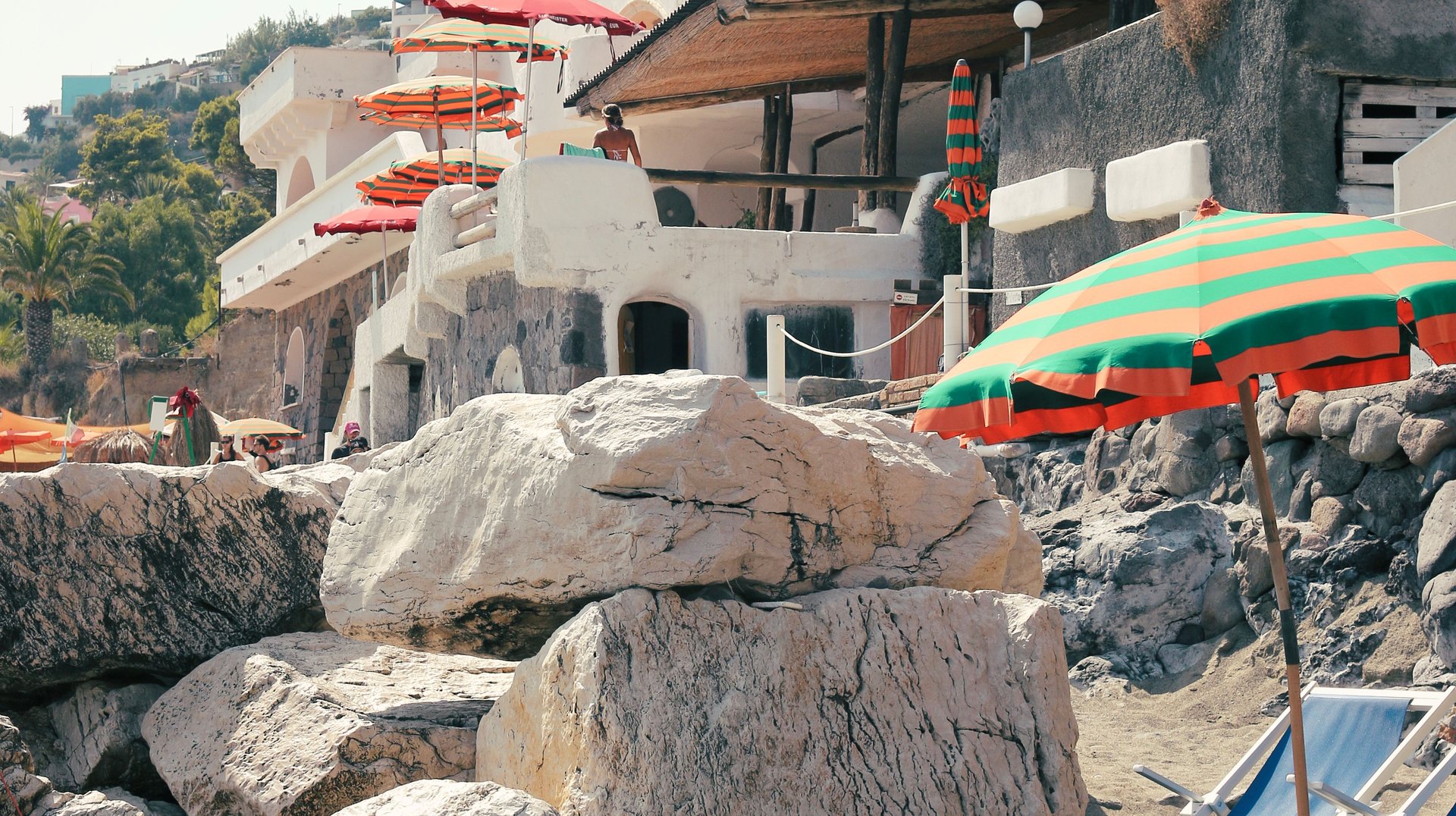 Coastal View Showcasing a Beach with Colorful Umbrellas, Lounge Chairs, and Rocky Shoreline Under Clear Skies.