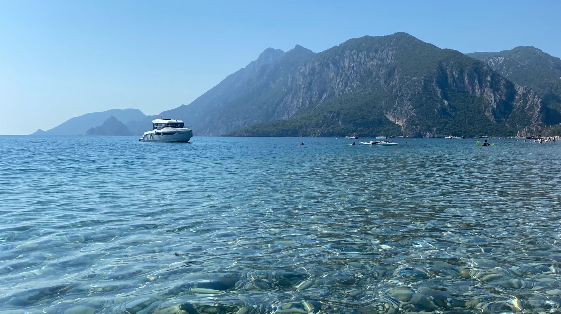Clear Waters and Mountainous Backdrop of a Mediterranean Shore Featuring a Distant Yacht.