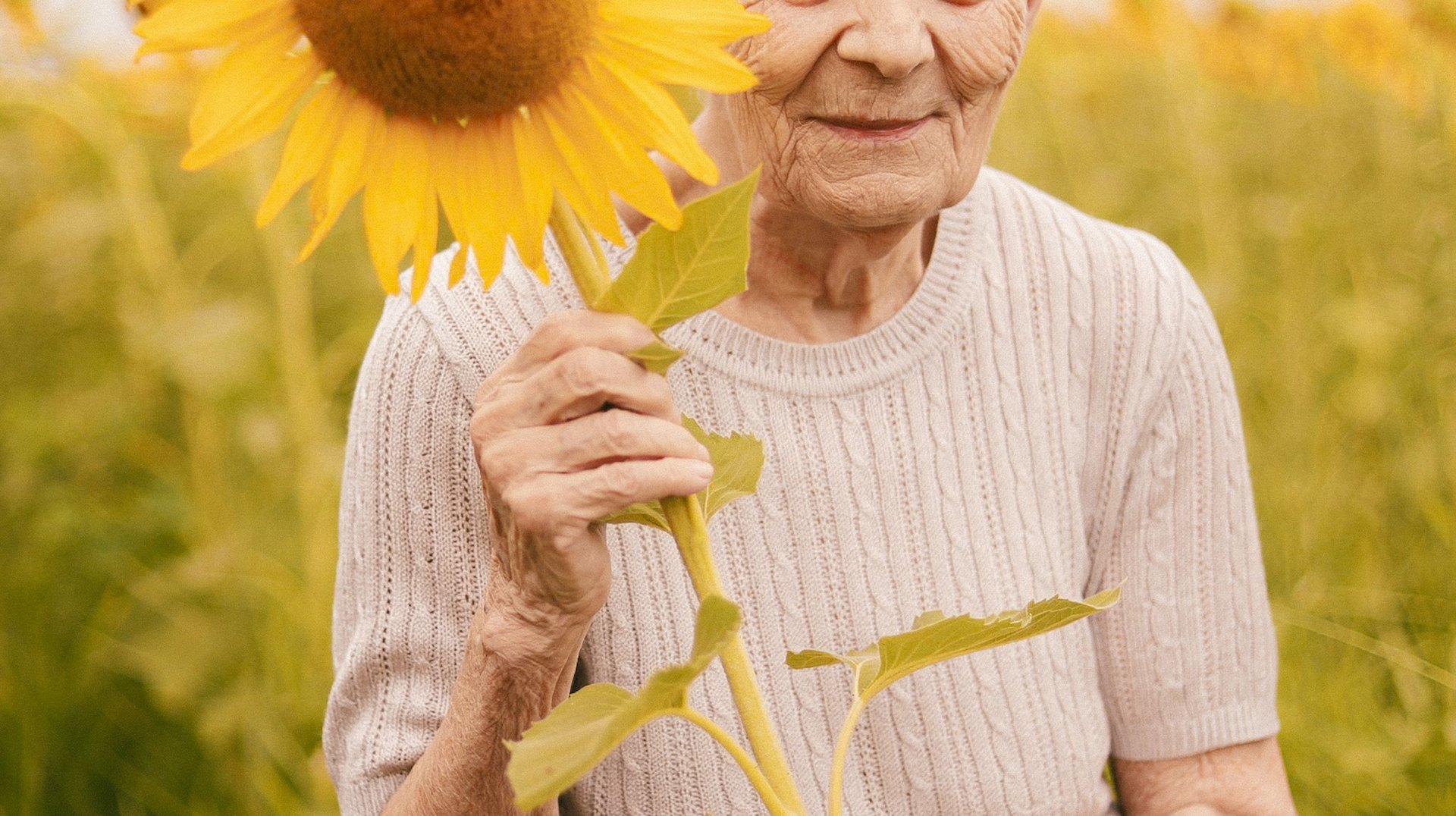 Senior Woman Joyfully Holding a Sunflower in a Vibrant Field During Summer, Expressing Warmth and Nostalgia.