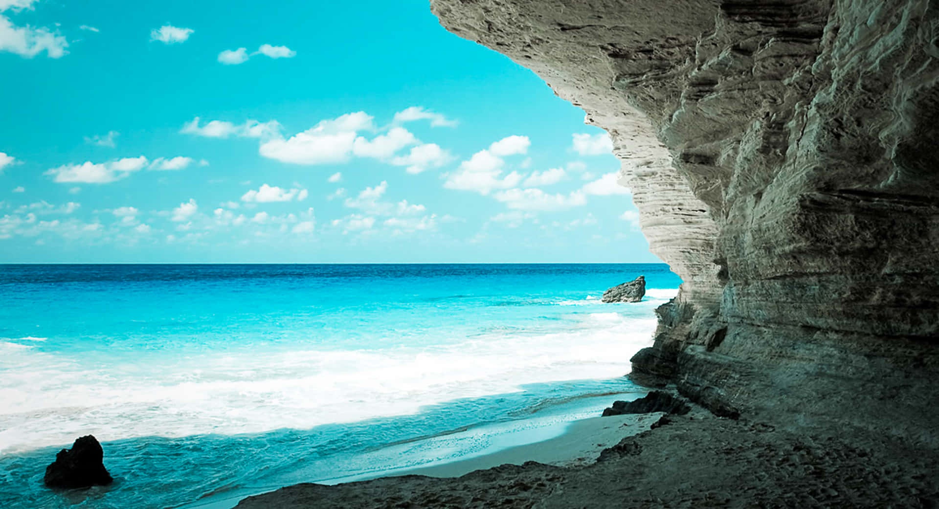 View from a cave opening to a turquoise ocean and sandy beach under a bright blue sky.