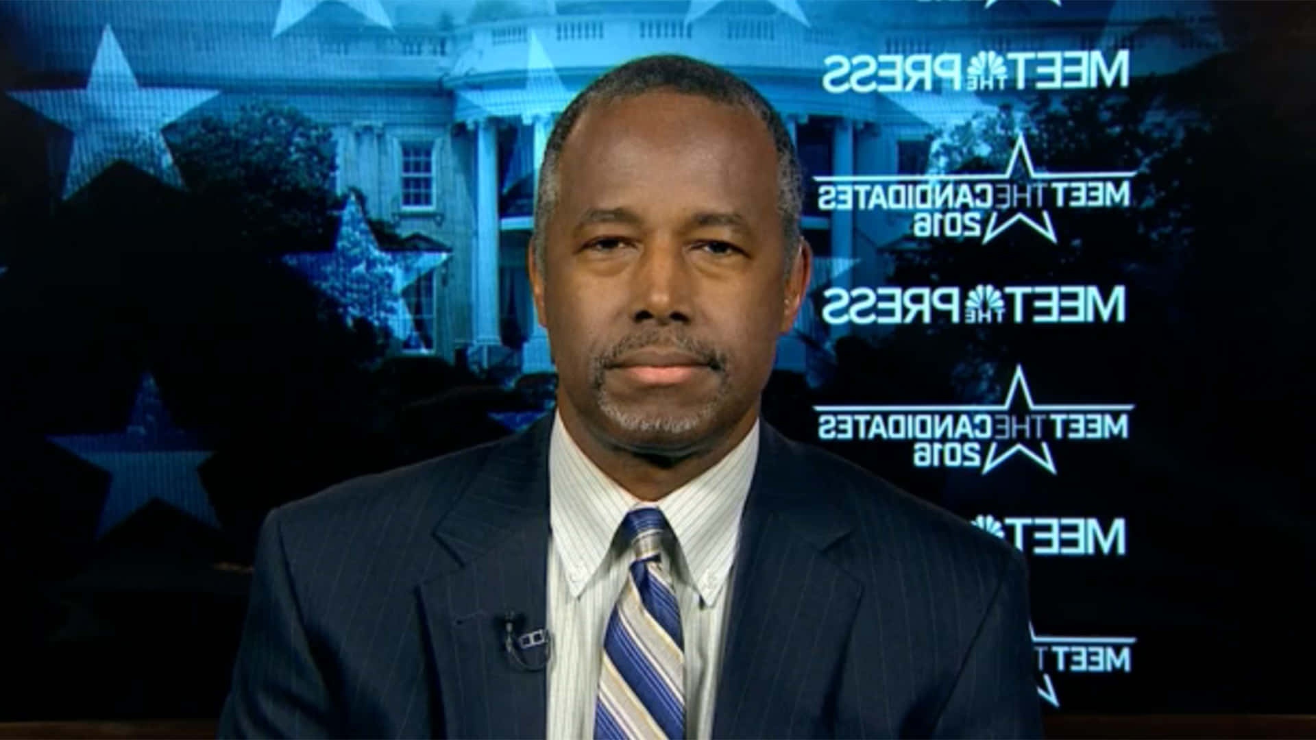 News anchor in suit and tie on 'Meet the Press' set with White House background.