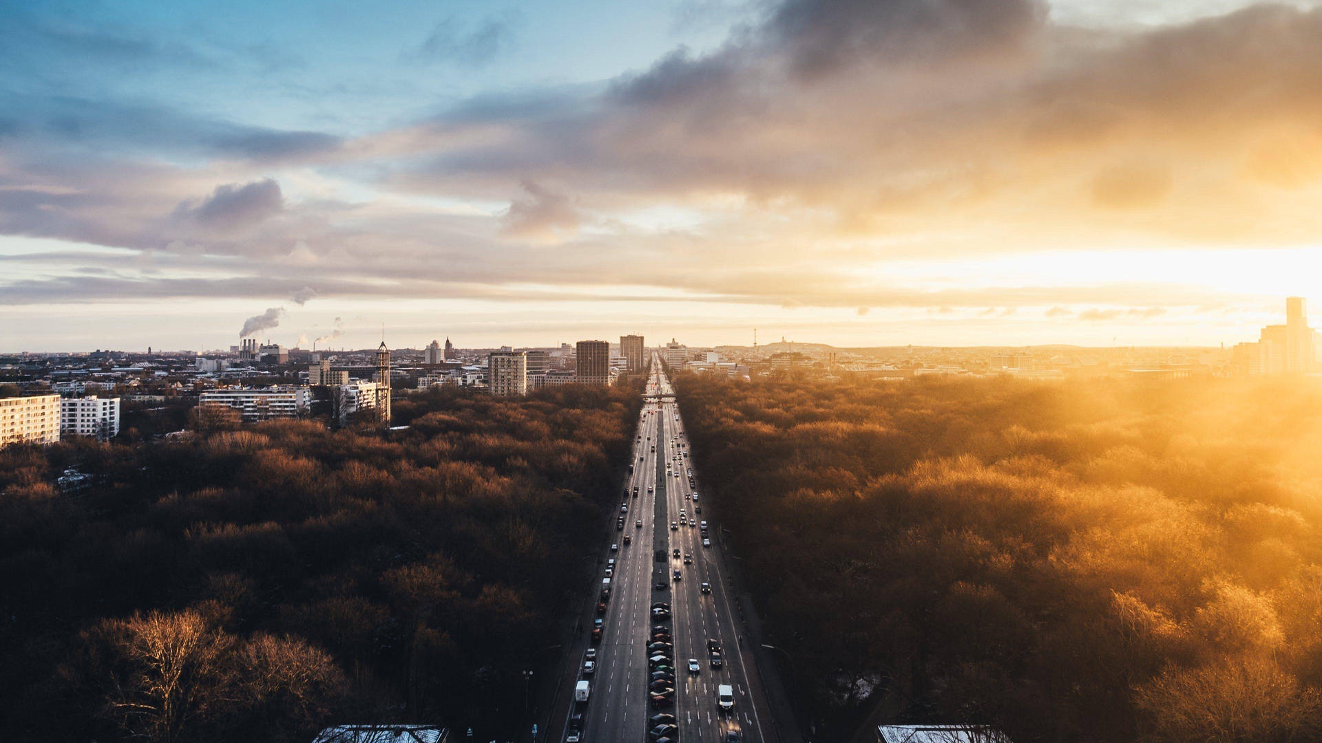 A highway through autumn trees at sunset with city buildings in the distance.