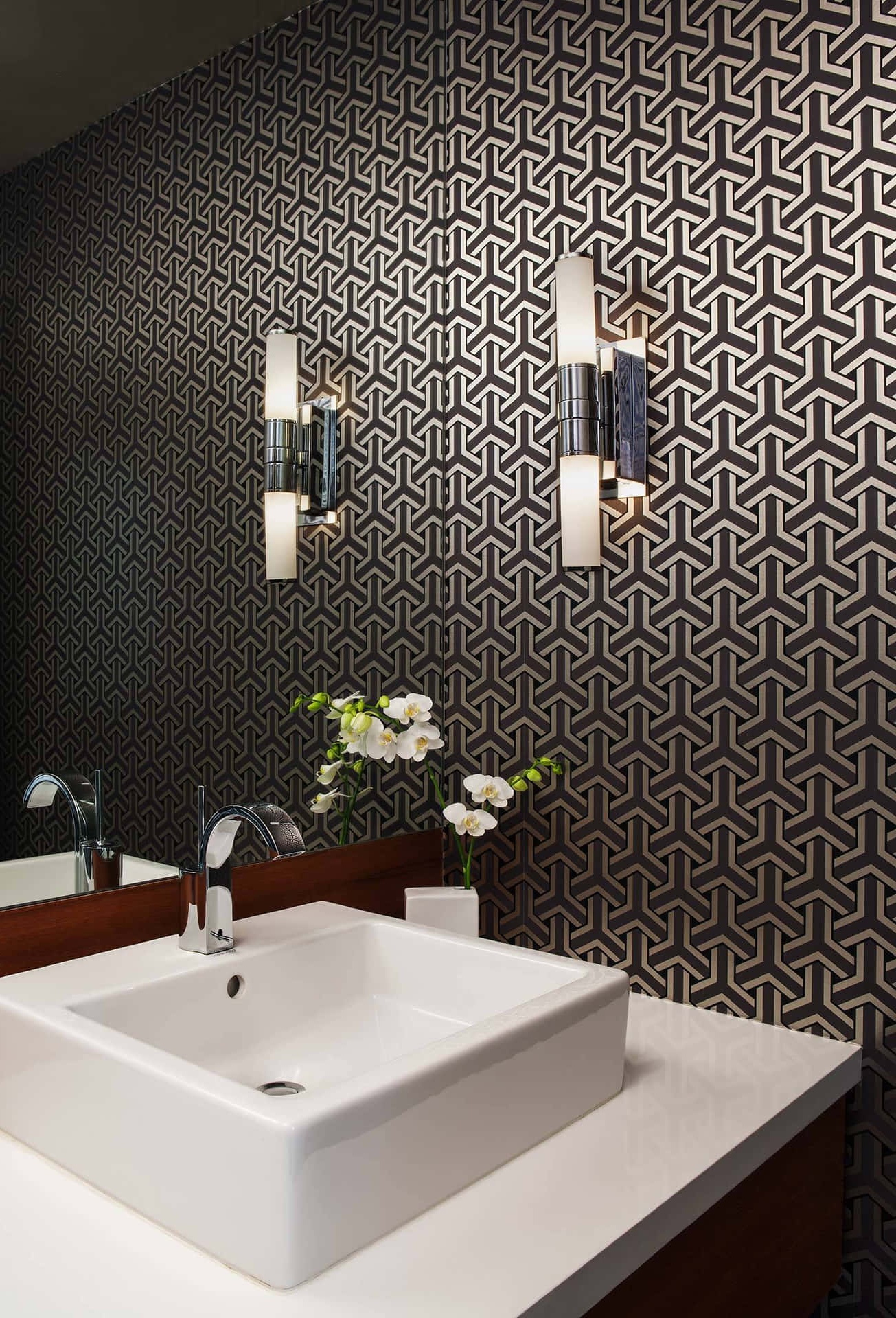 Modern bathroom with geometric wallpaper, white sink, and orchids under warm sconce lighting. – Ultra HD Wallpaper