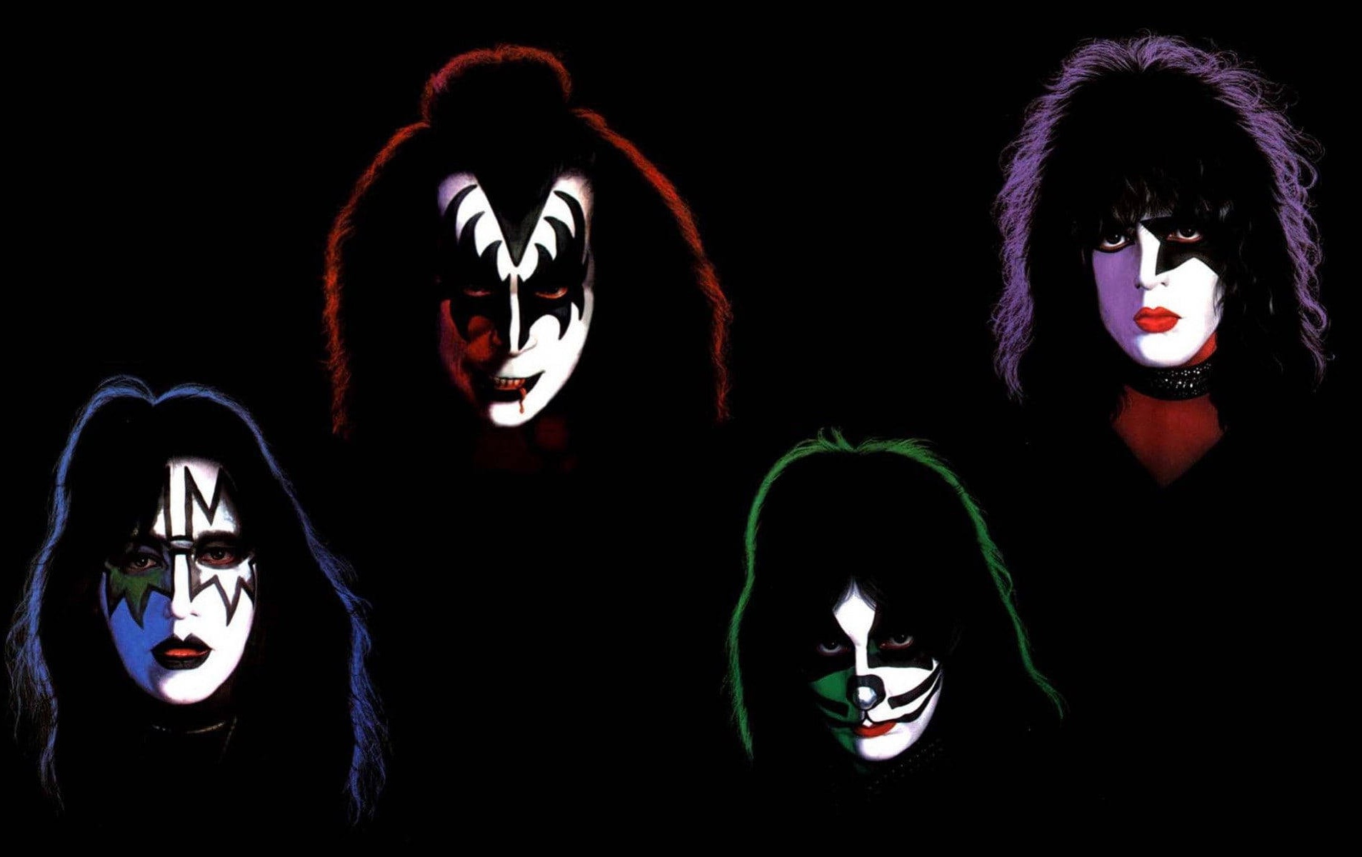 Four glam rock musicians with dramatic makeup and colorful hair against black.