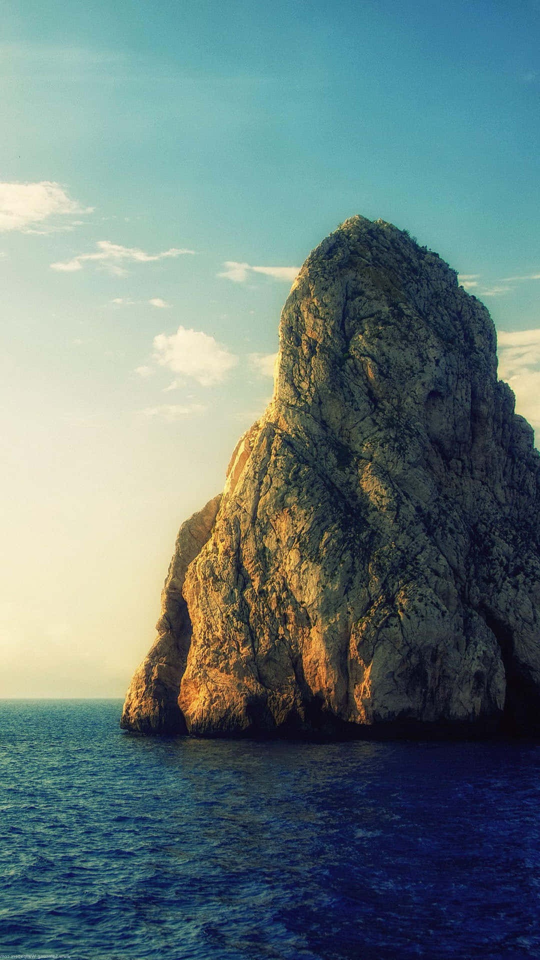 A large rocky island stands in the ocean at sunset with warm light on its cliffs.