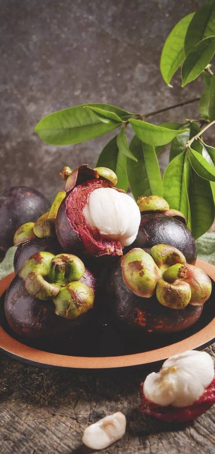 Ripe mangosteens in a bowl with green leaves on a wooden surface.
