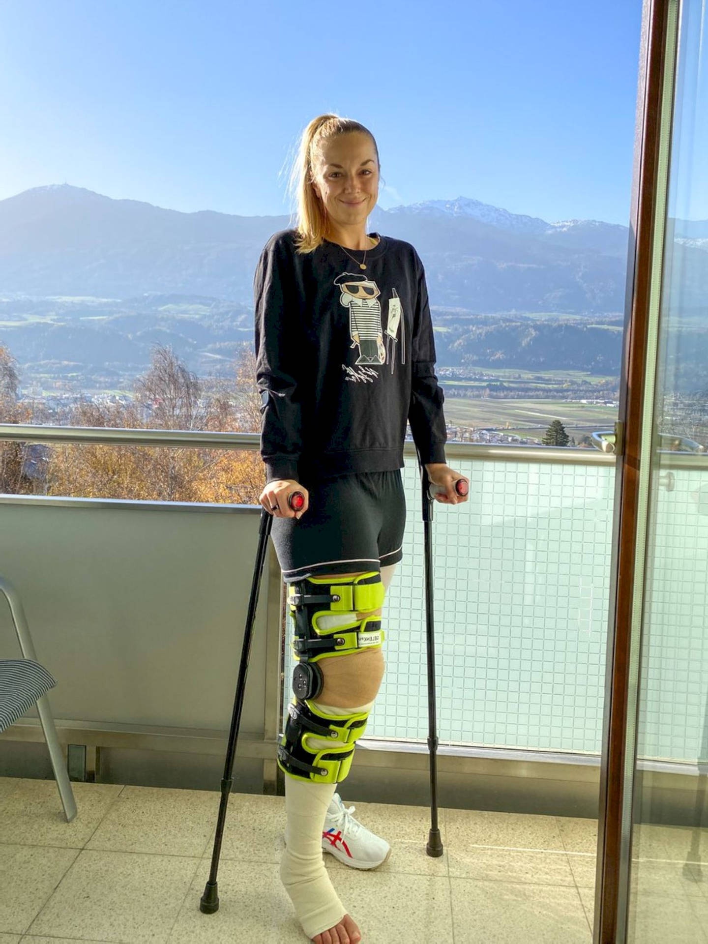 Woman with knee brace and crutches smiling on a mountain-view balcony.