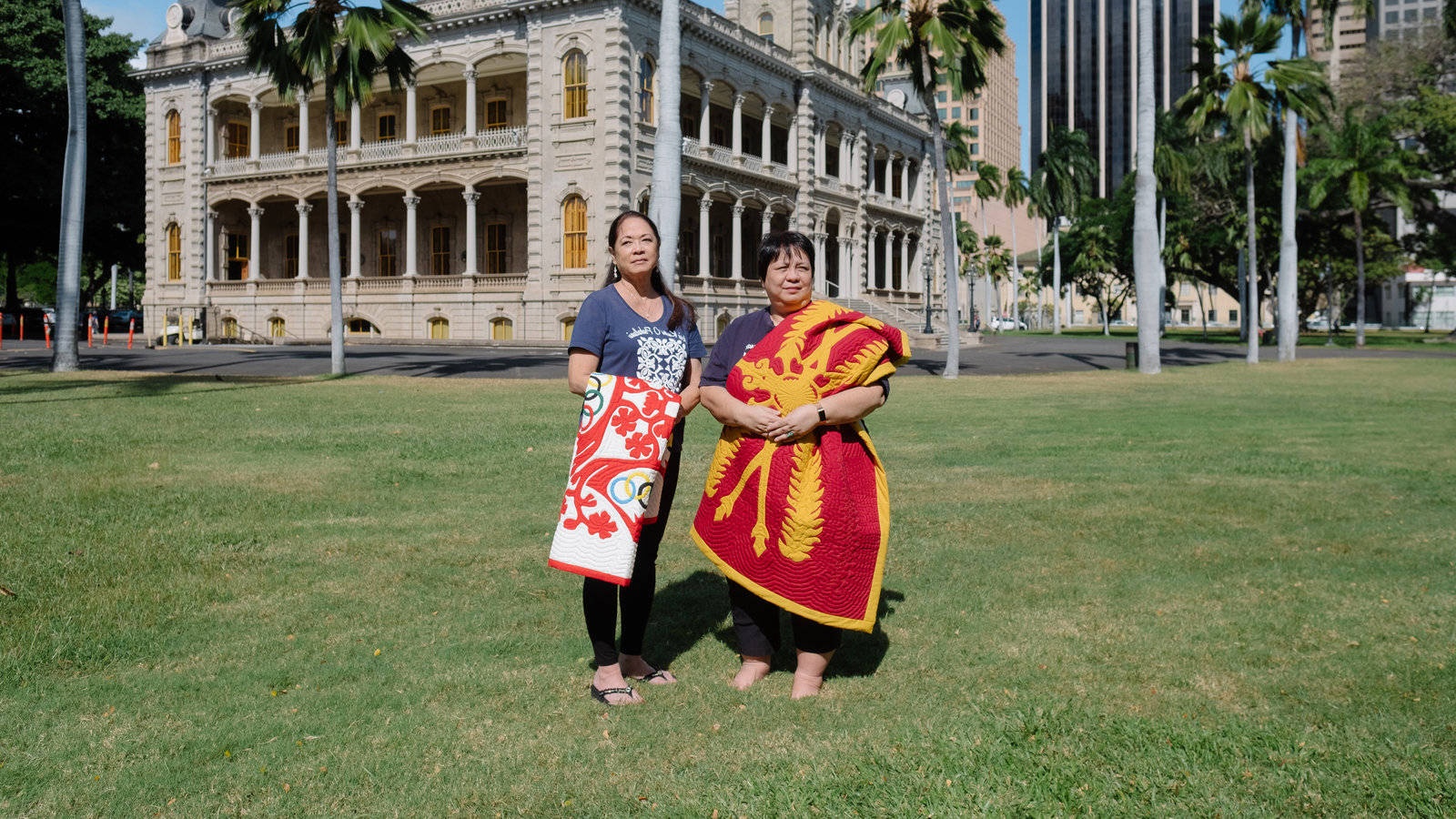 Two women holding patterned blankets in front of a historic building with palm trees. – Ultra HD Wallpaper