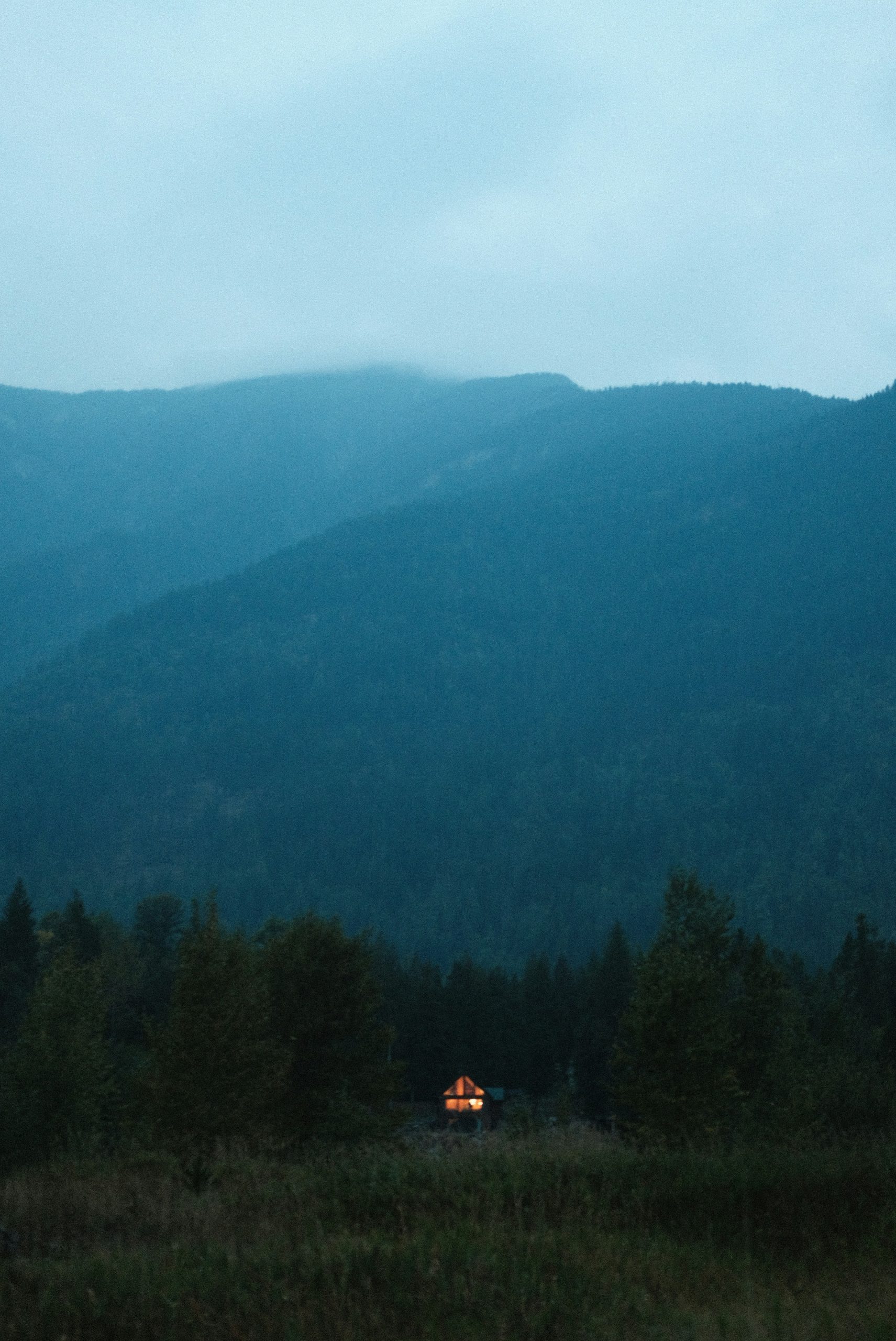 Small cabin glows in the twilight forest before mountains.