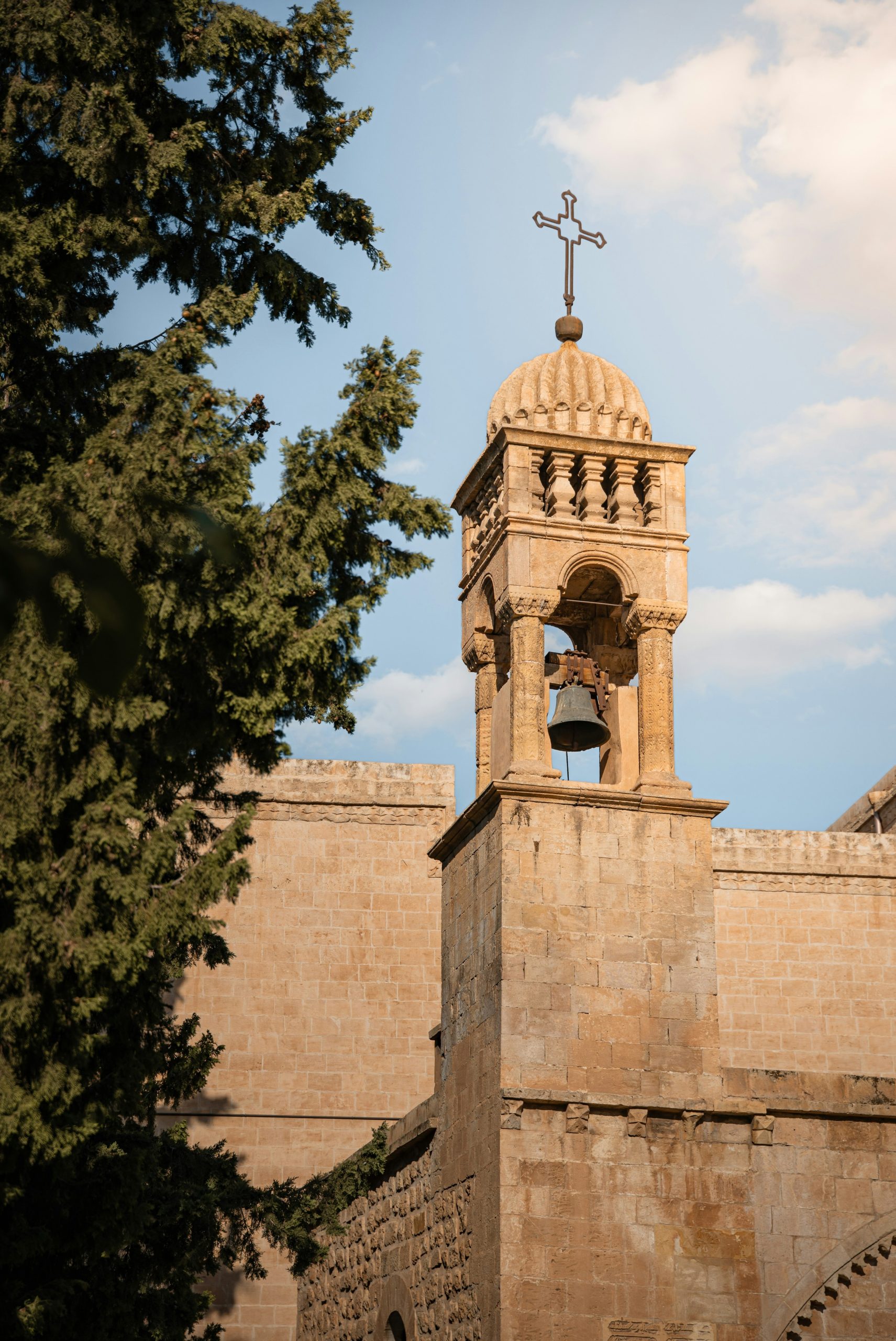 Stone bell tower with cross and bell against sky.