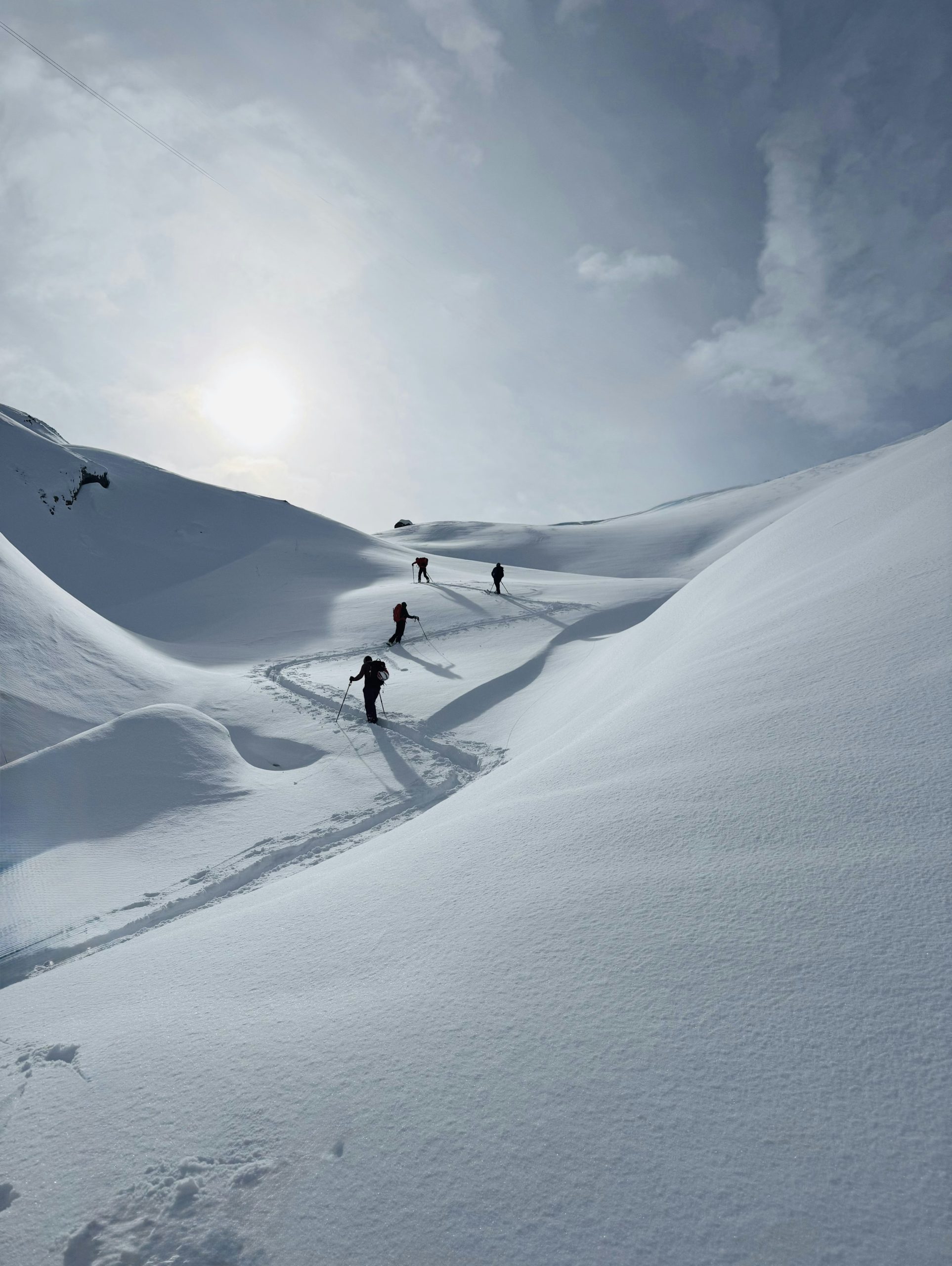 Skiers ascend a snowy mountain path under a bright sun.