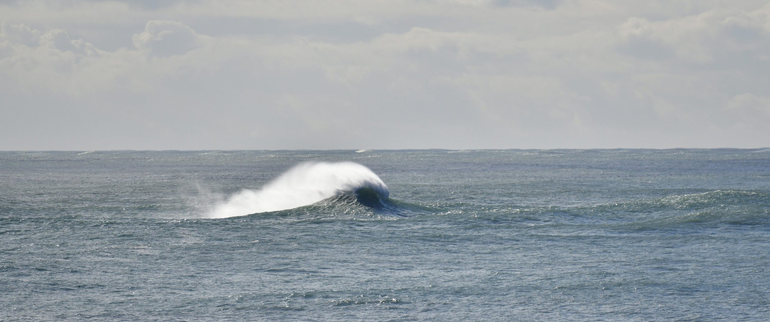 A wave crests in the ocean under a cloudy sky.