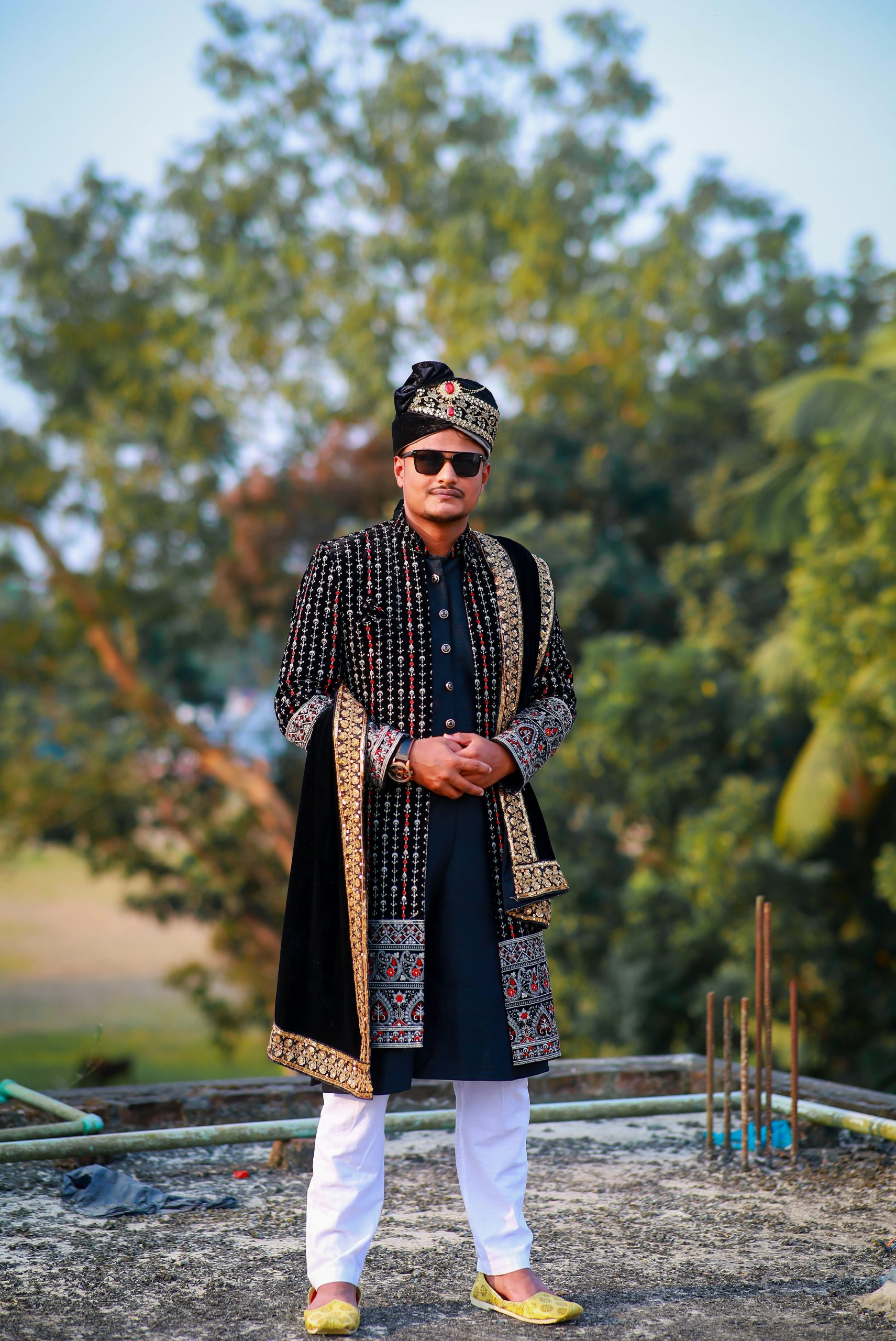 A man in traditional sherwani and turban stands outdoors.