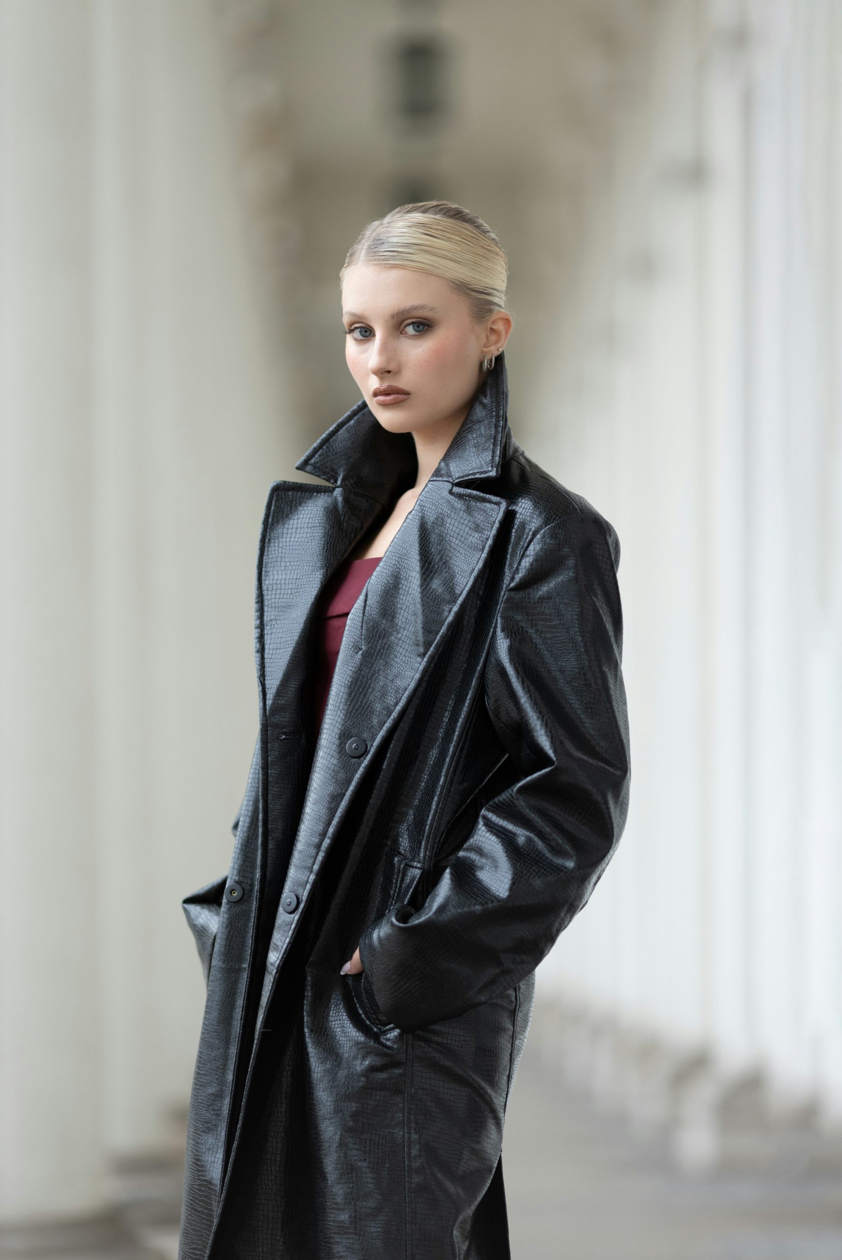 A young woman in a black trench coat stands outdoors.