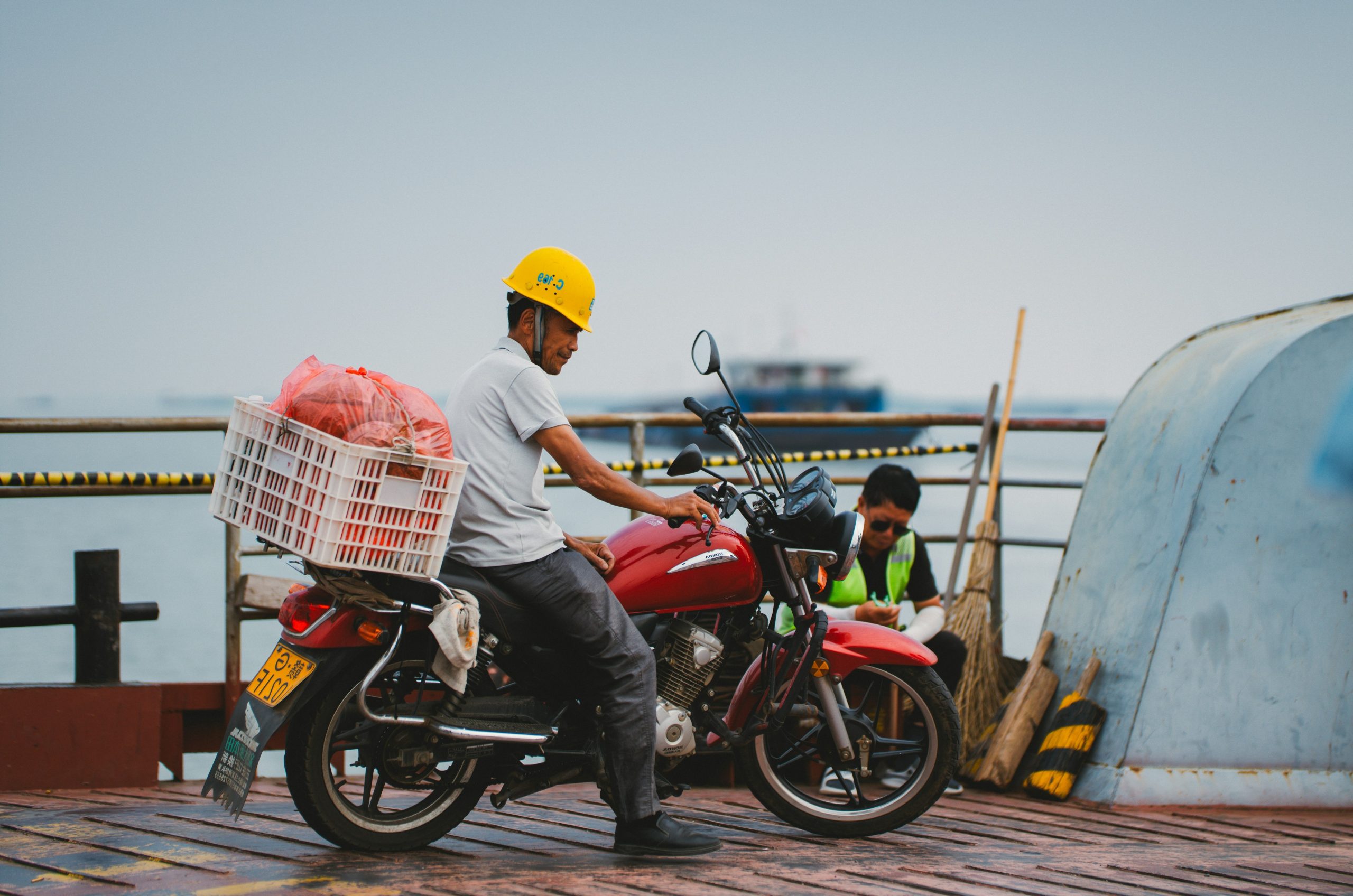 Man on red motorcycle with goods near water.