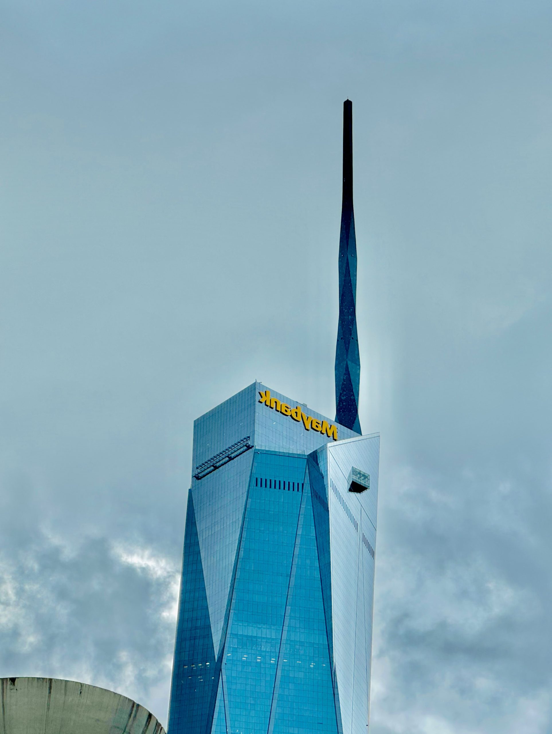 Maybank skyscraper with a tall spire under a cloudy sky.