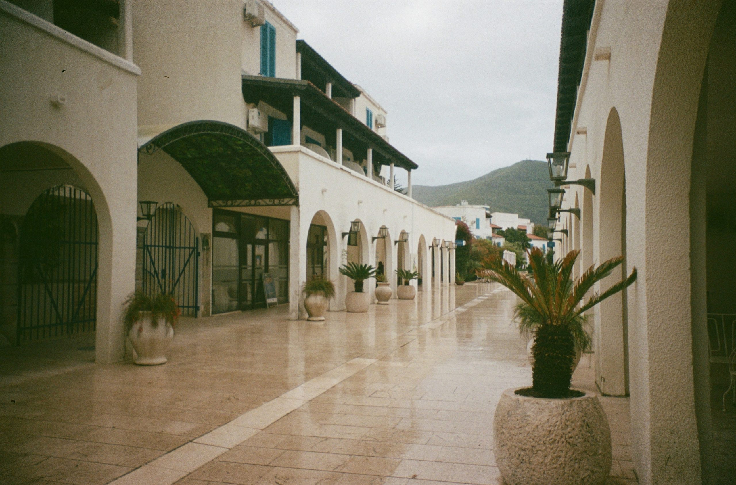 White arched buildings line a wet paved walkway with mountains.