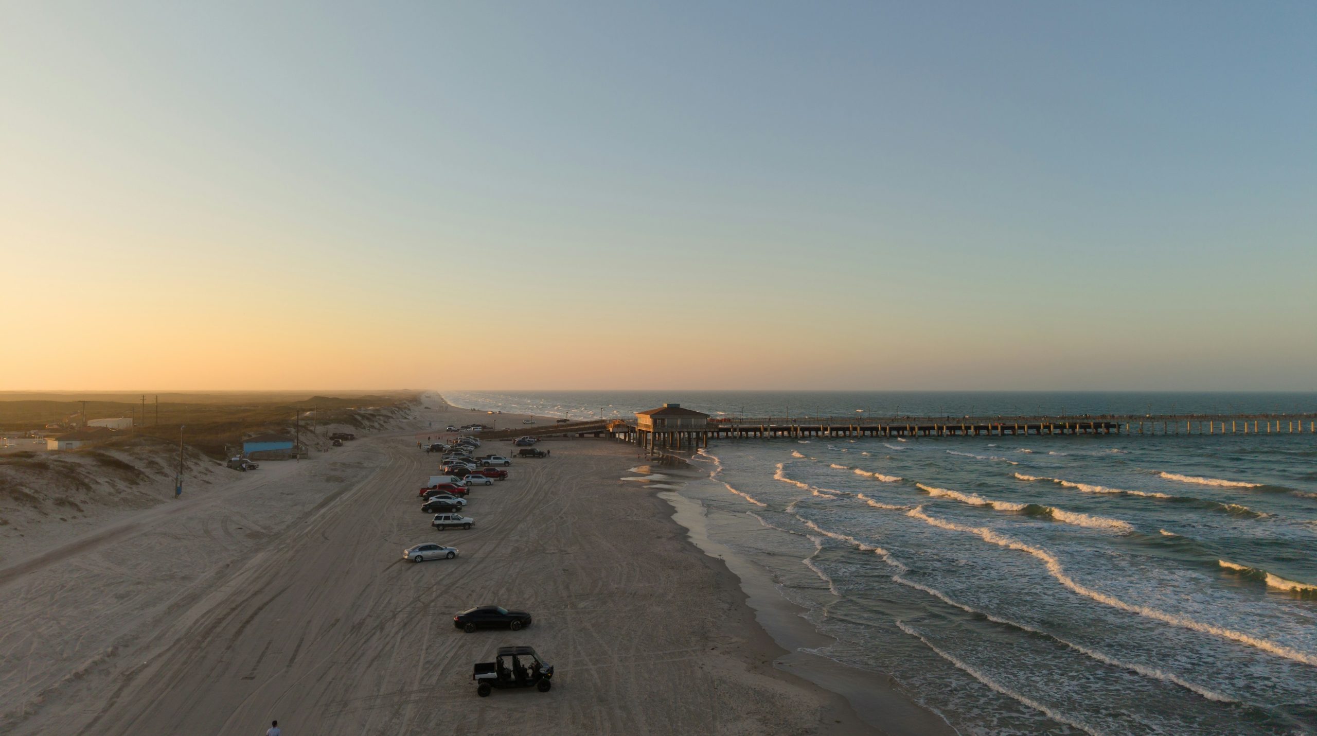 Coastal beach with pier, ocean waves, and cars at sunset.
