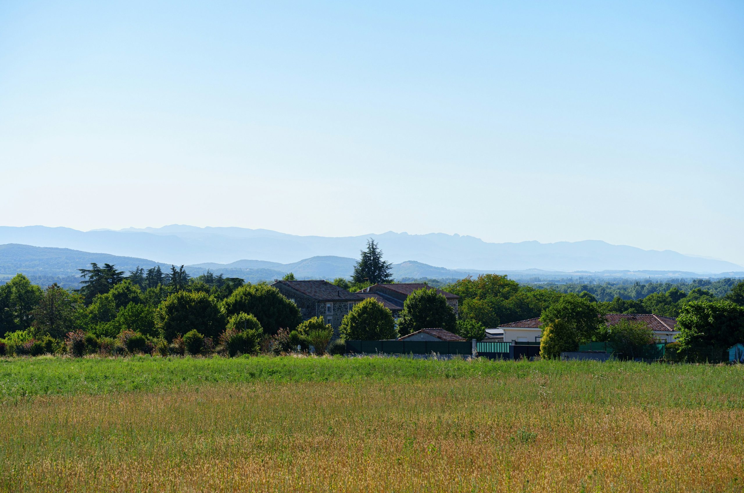 Houses among trees, dry field, distant mountains, clear blue sky.