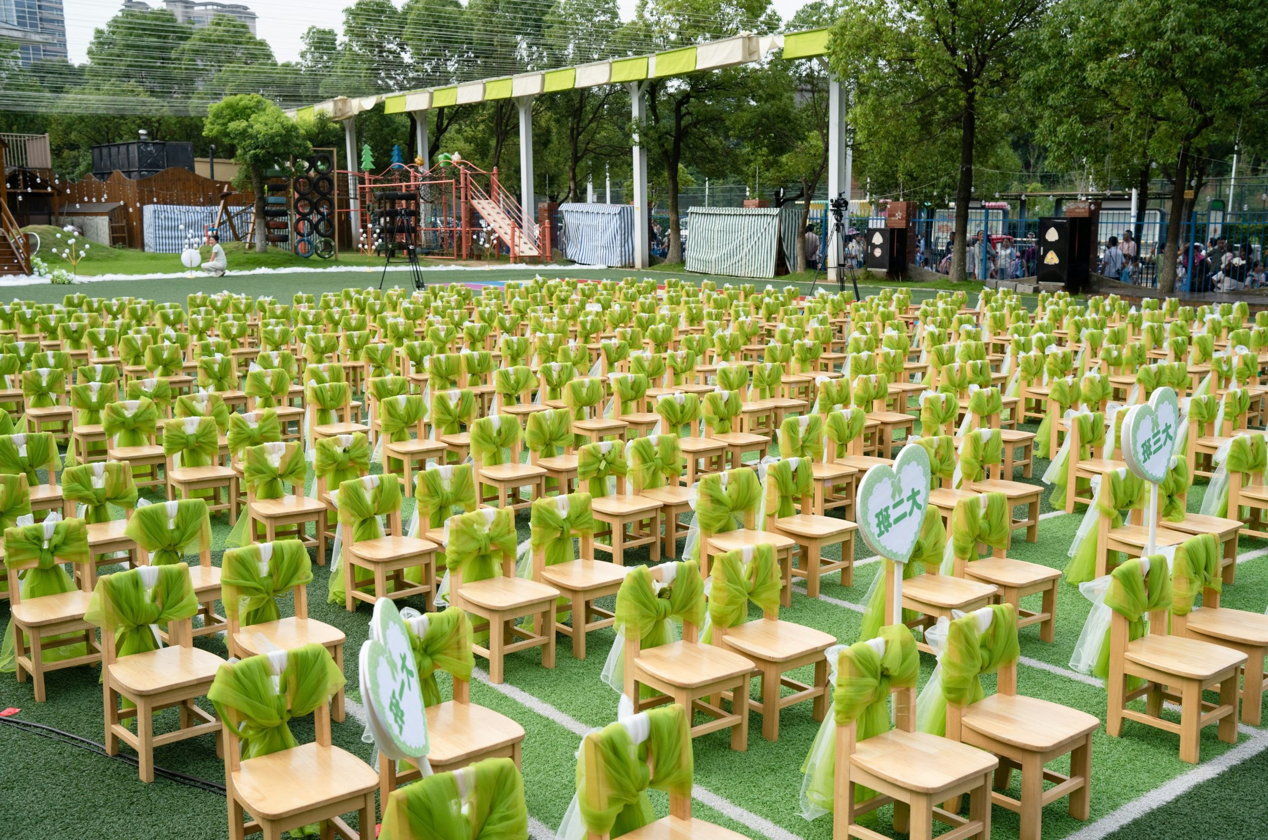 Many small wooden chairs with green sashes on artificial turf.