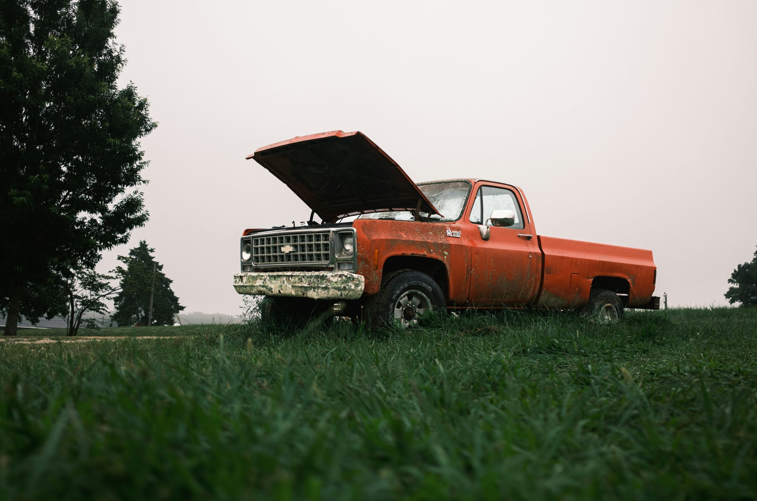 Rusty orange pickup truck with hood open in a field.