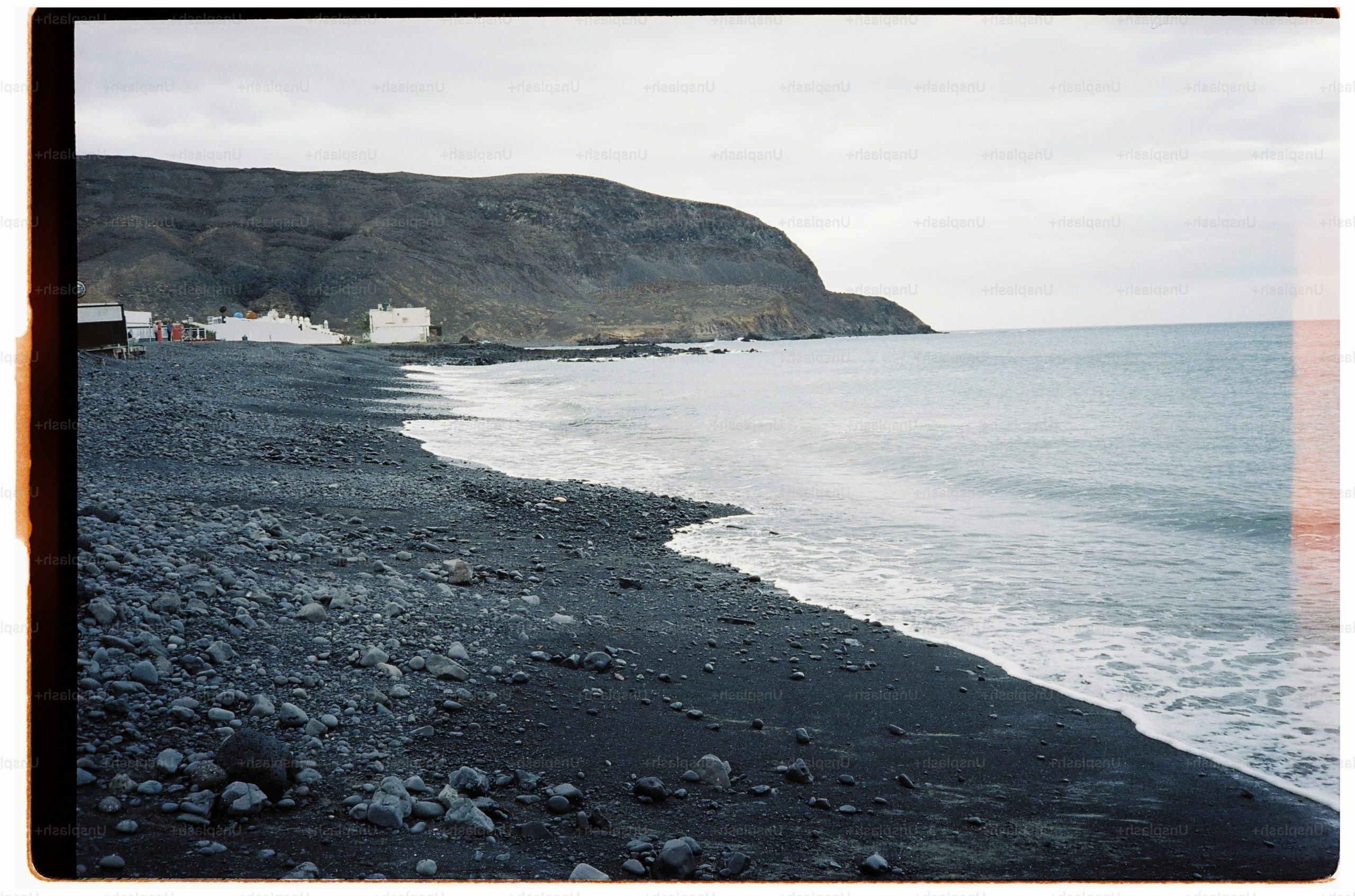Waves washing onto a dark volcanic pebble beach beneath a large coastal cliff.