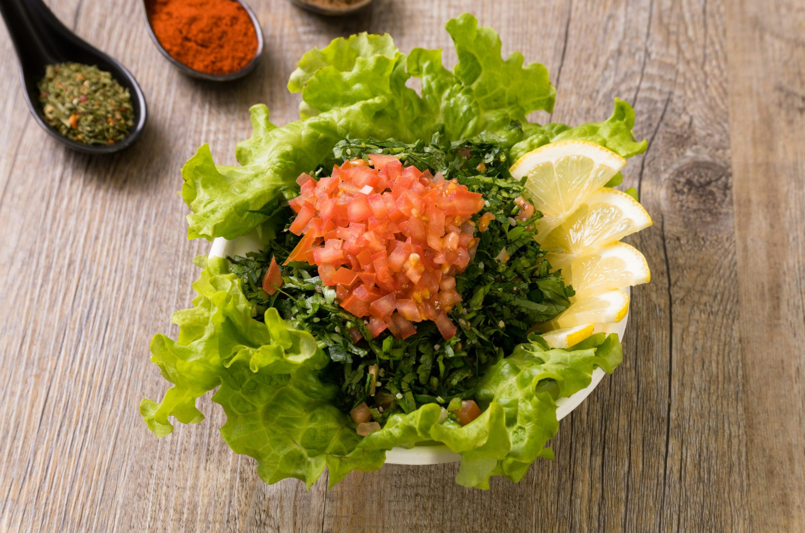 A bowl of tabbouleh salad with chopped tomatoes, parsley, and lemon slices on wood.