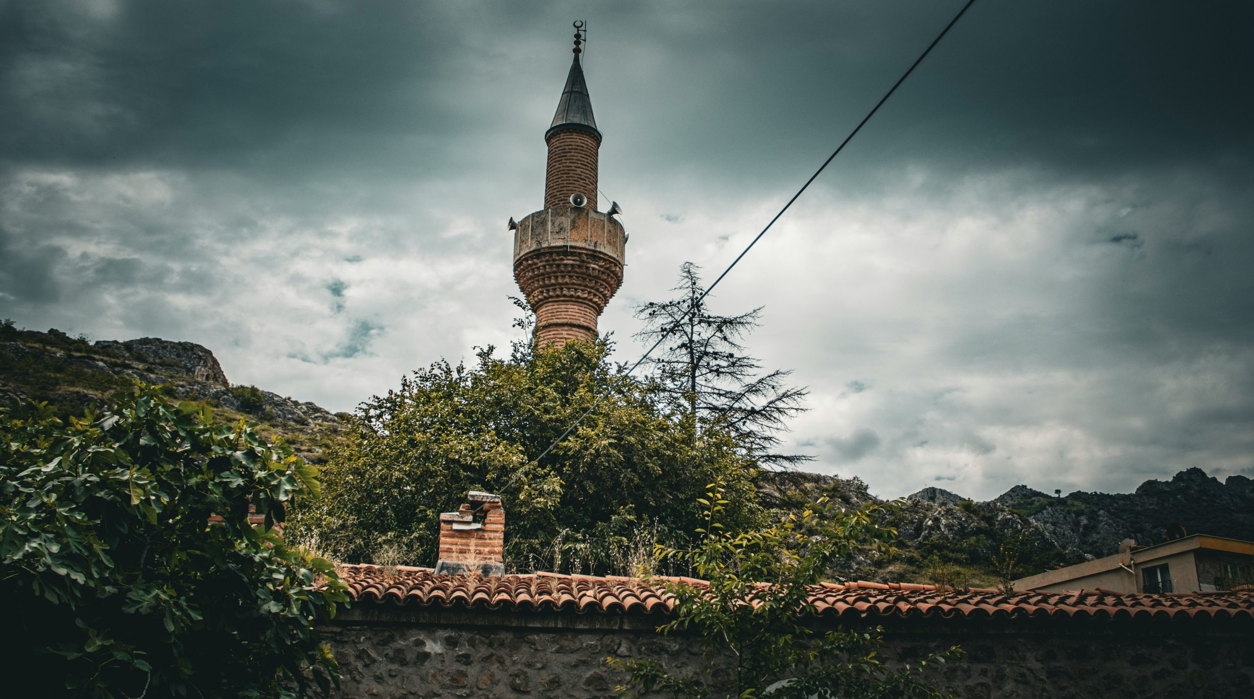 A brick minaret rising above trees and a tiled roof under a cloudy sky.