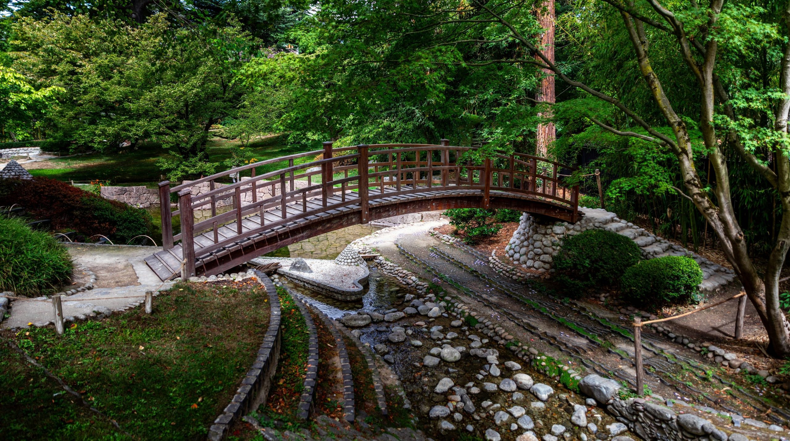 A wooden arched bridge over a rocky stream in a lush green garden.