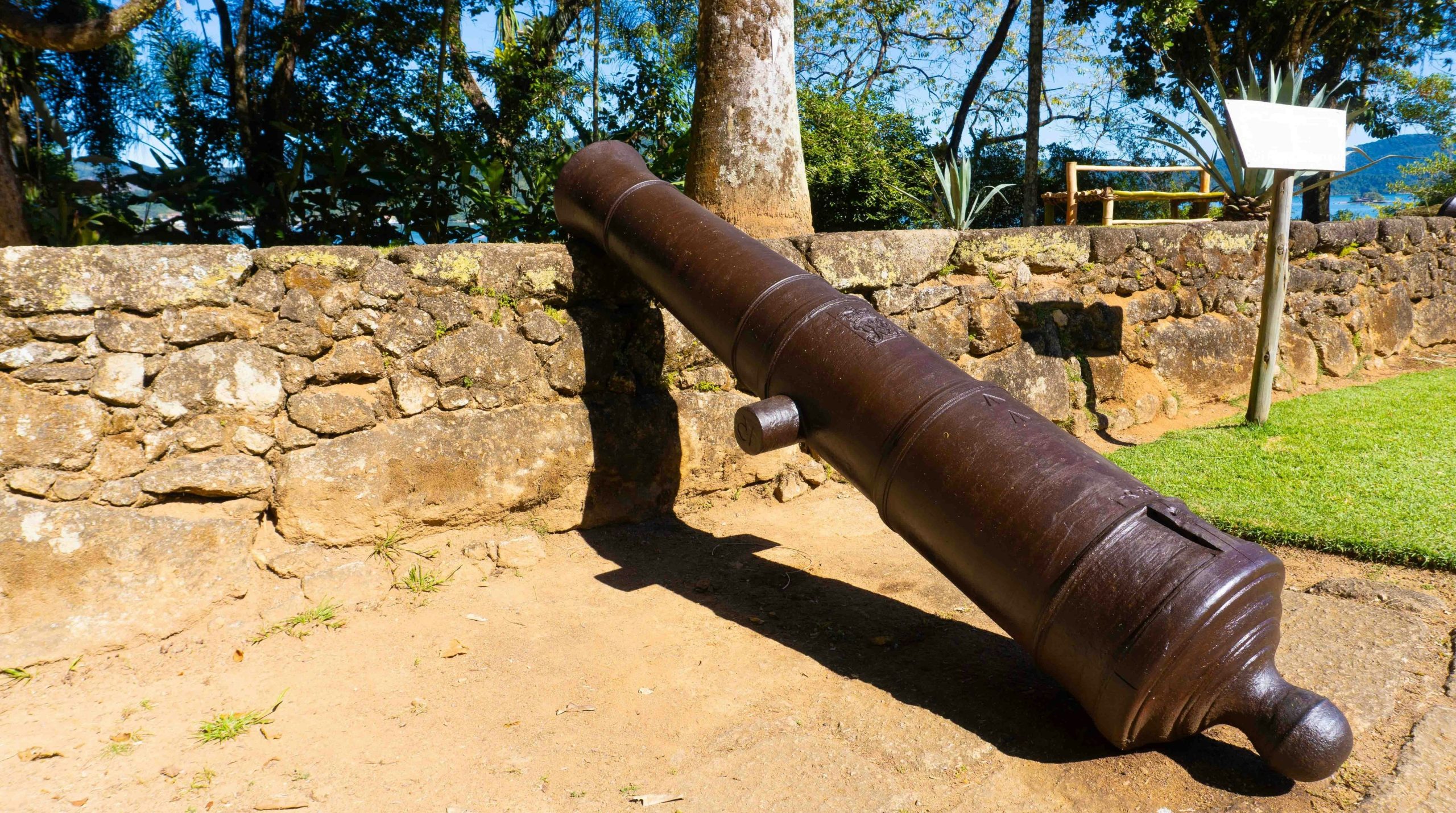 A dark metal cannon resting against a stone wall in a sunny garden.