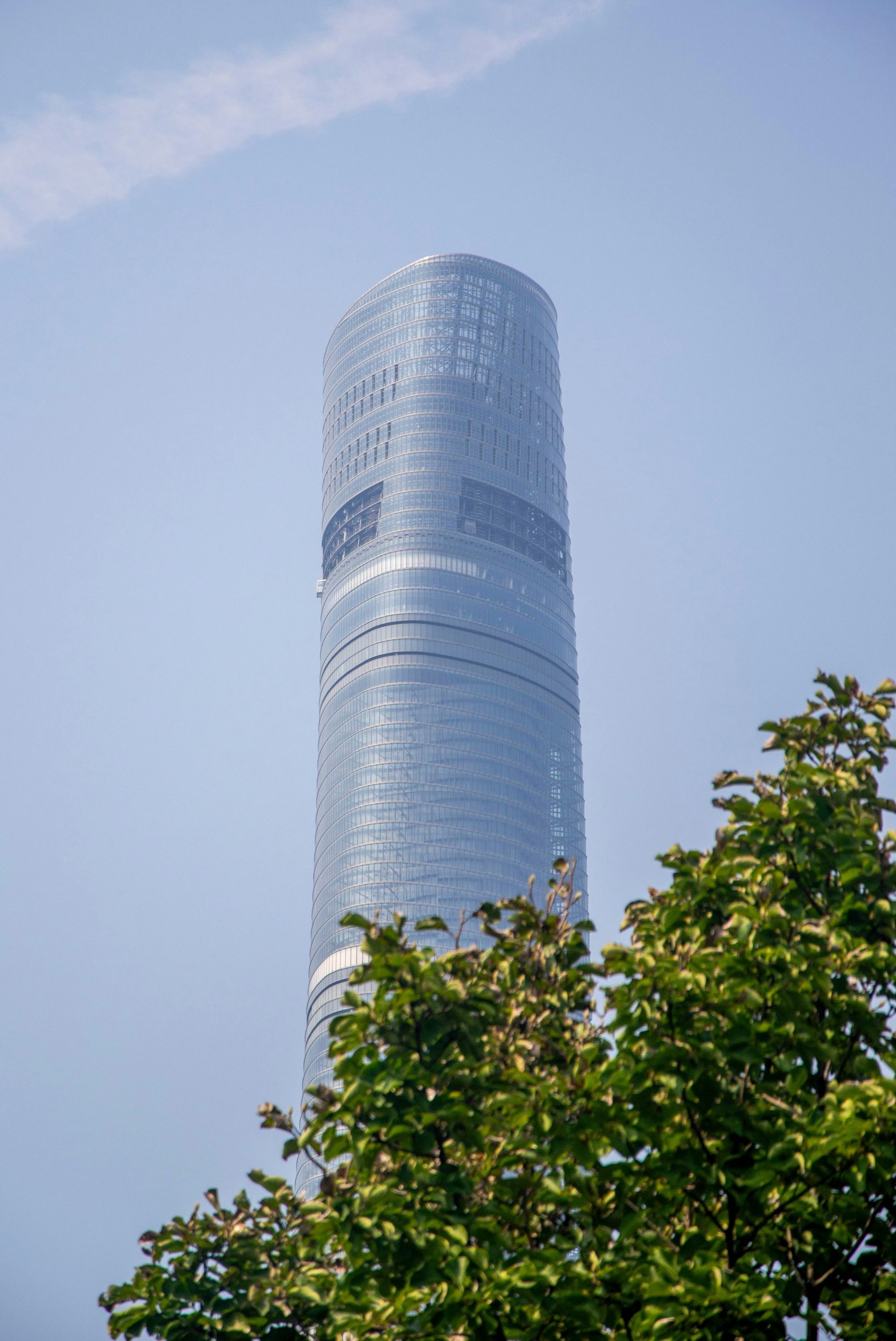 A tall cylindrical glass skyscraper rising behind green tree foliage against a clear sky.