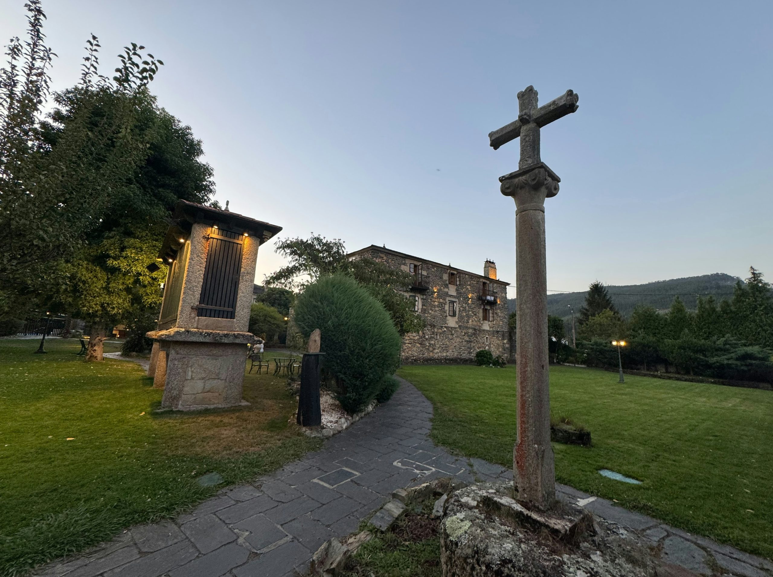 A stone cross on a pedestal in a garden with a stone granary.