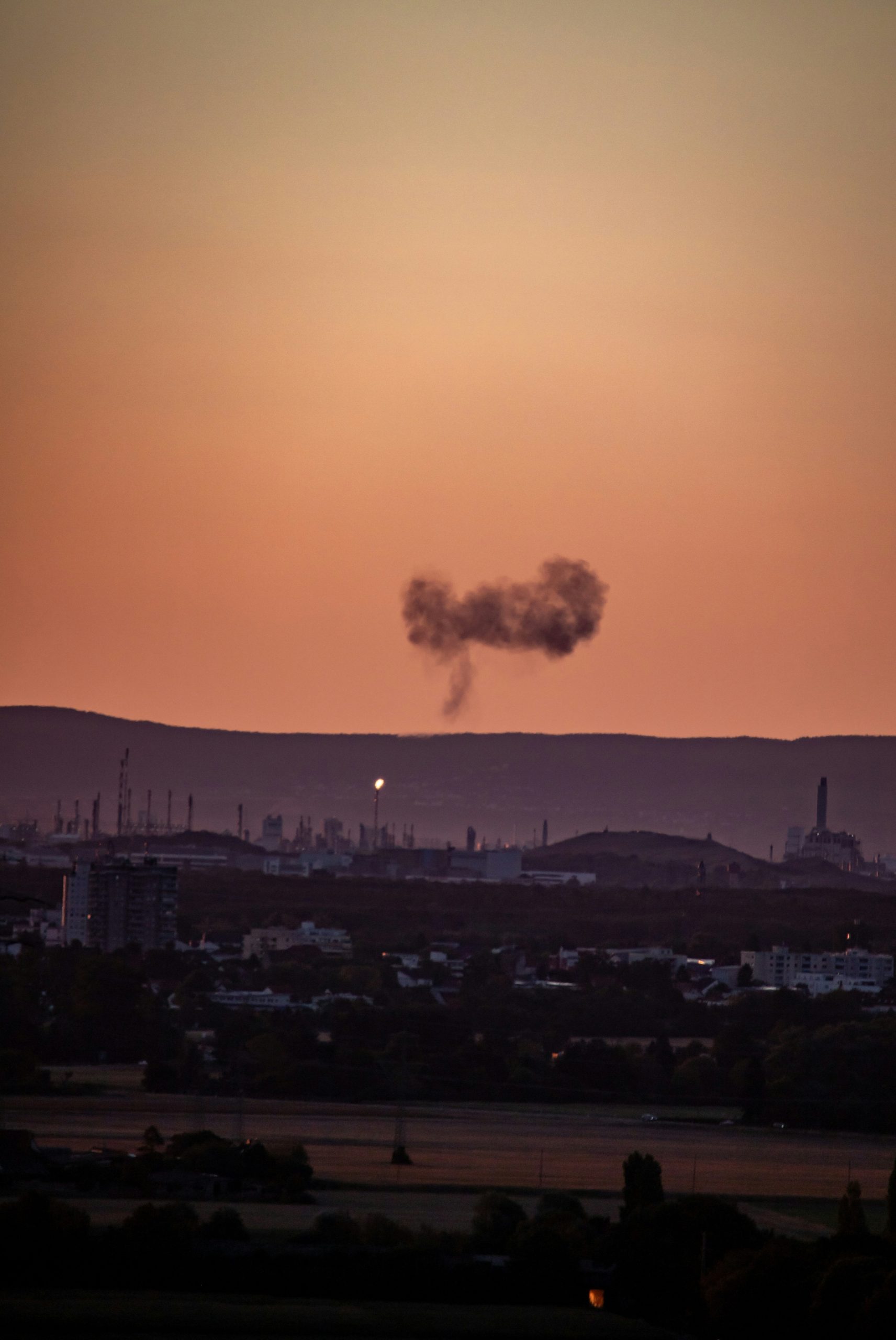 A dark plume of smoke rising above an industrial skyline at golden hour.