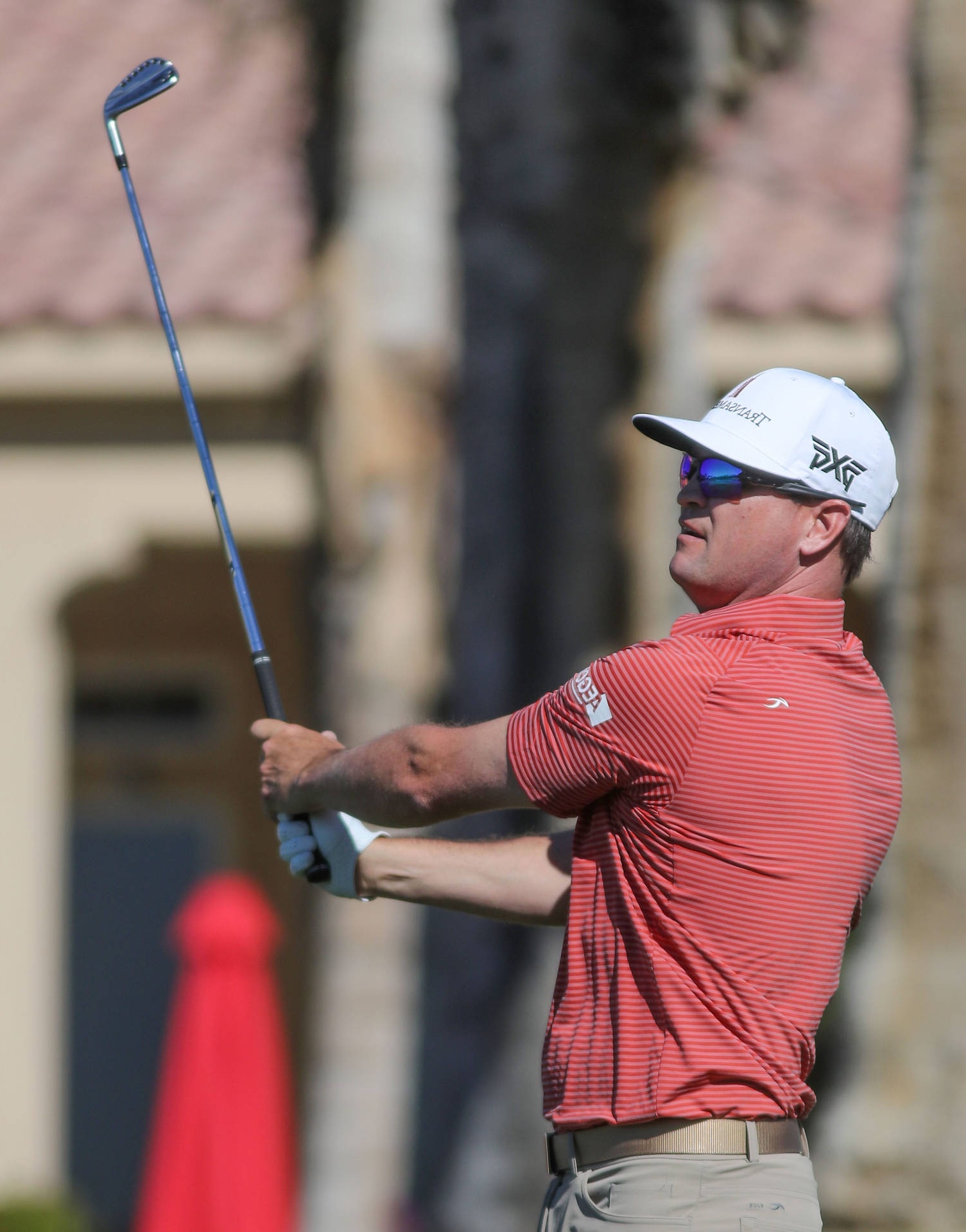 Golfer mid-swing wearing red shirt and white cap on sunny day.